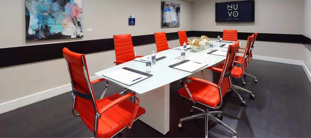 Sala de reuniões com mesa branca, dez cadeiras cor de pêssego, quadros nas paredes e uma televisão de tela plana